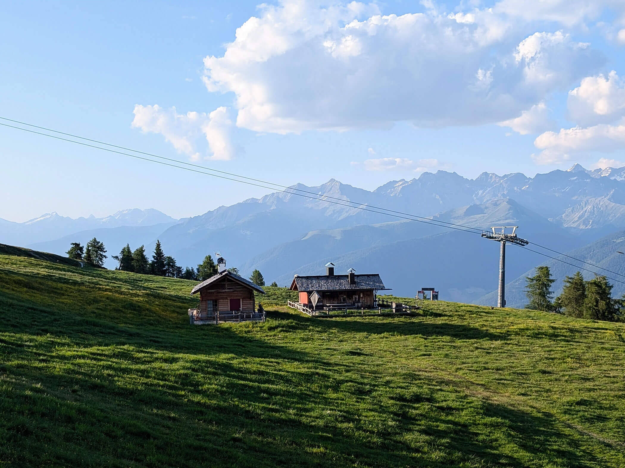 Zwei Almhütten auf einer grünen Wiese und dahinter eine Seilbahn - Branterhof